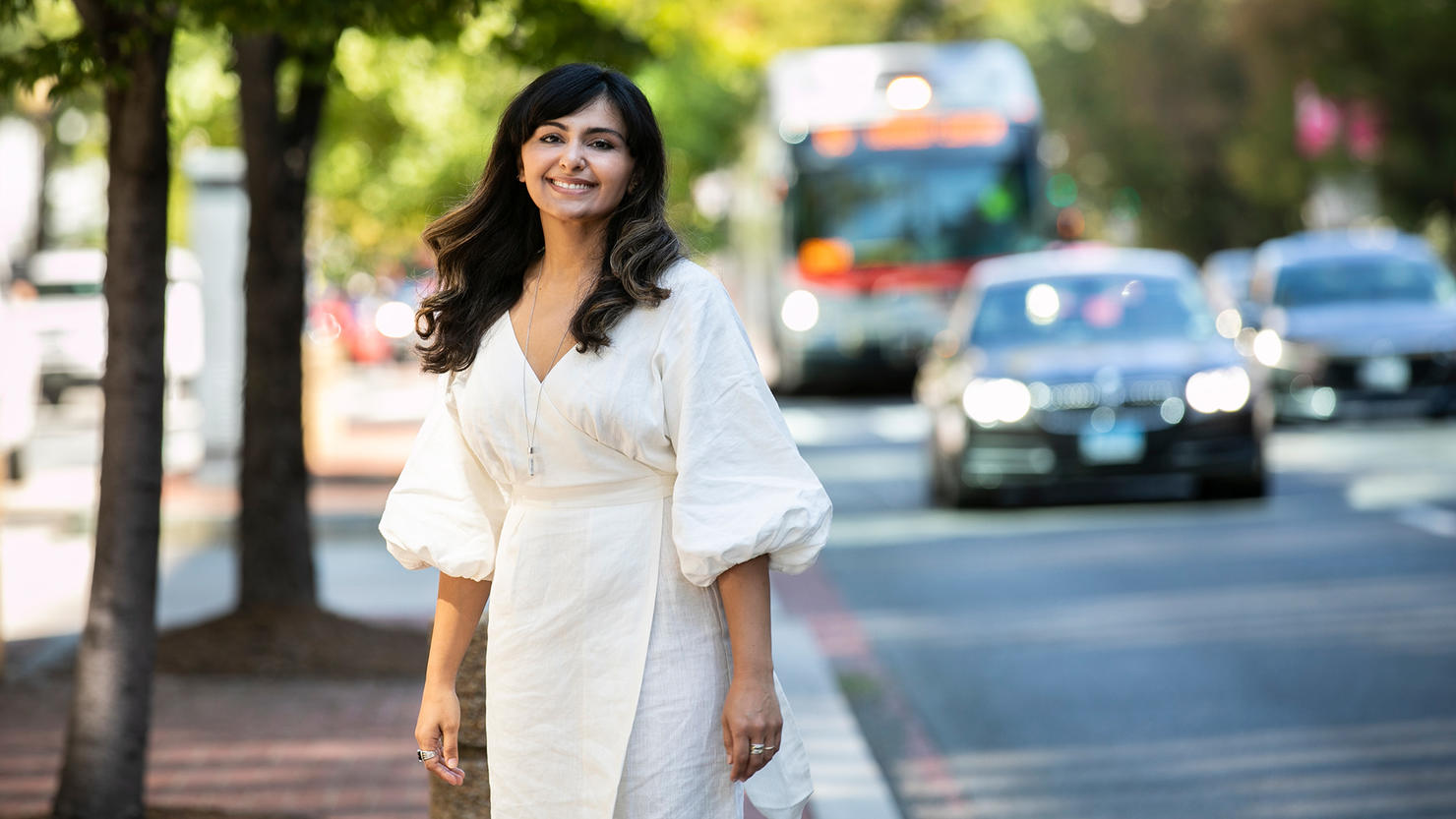 Dilafruz Khonikboyeva standing outside on the sidewalk wearing a white dress. She is smiling at the camera.