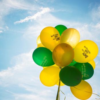 A bunch of green and gold balloons float against a bright blue sky