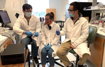 Three researchers in lab coats and masks sit in a lab-type setting